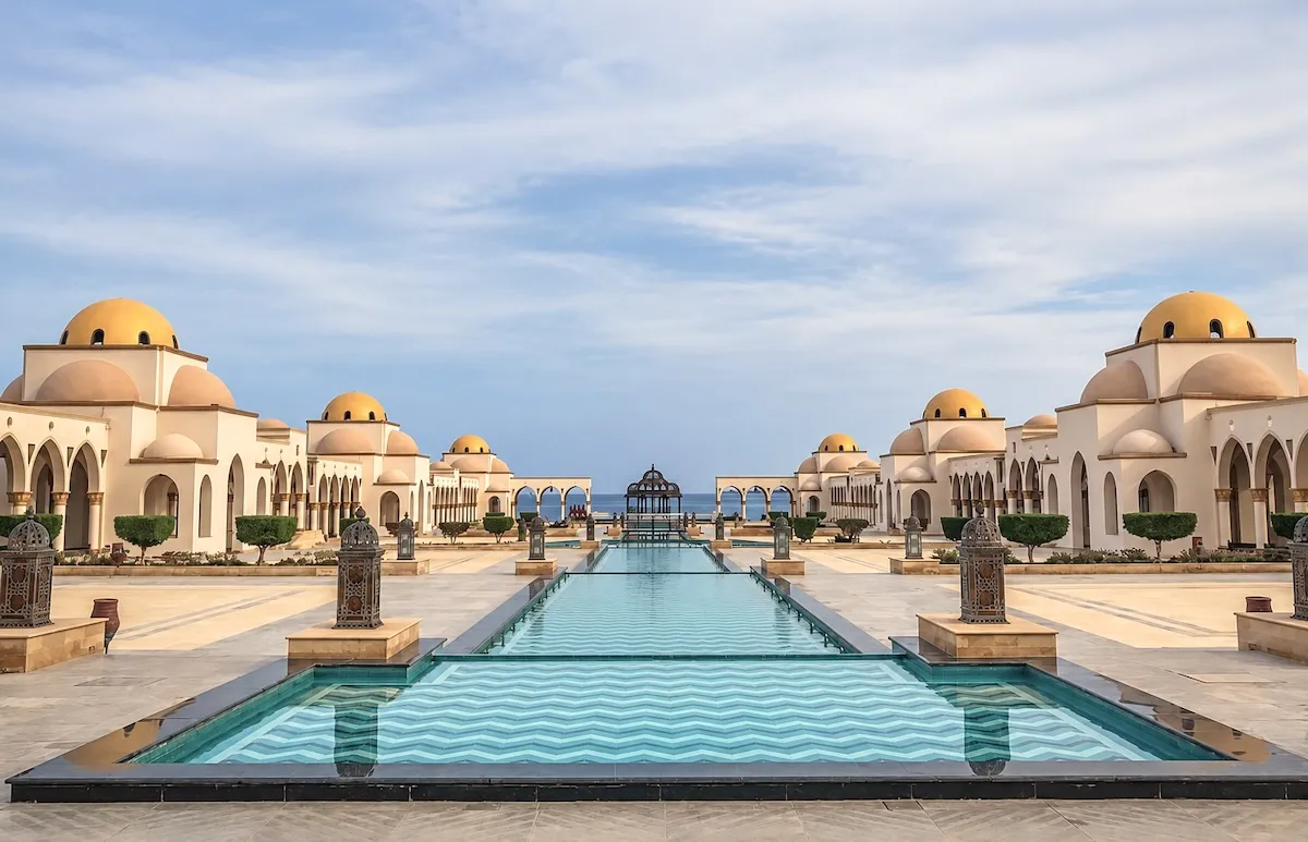 Domed white buildings and arcades on either side of a long turquoise water basin leading to the sea, with lanterns along the edges