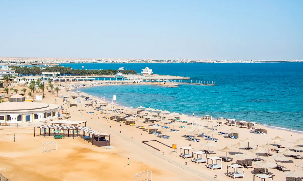 Ein Hotelstrand in Sahl Hasheesh mit Strohsonnenschirmen, Sonnenliegen und weißen Strandhütten entlang einer türkisfarbenen Bucht sowie einem Pier in der Ferne